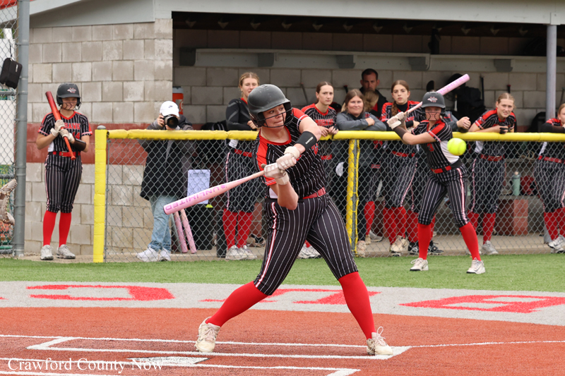 Female softball batter in a red-and-black uniform swings at a pitch on a red dirt field with a yellow fence and dugout in the background.