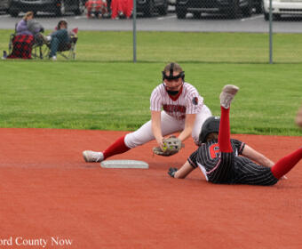 Softball infielder in white and red reaches for the ball near second base as a runner slides into the bag on a red-dirt field.