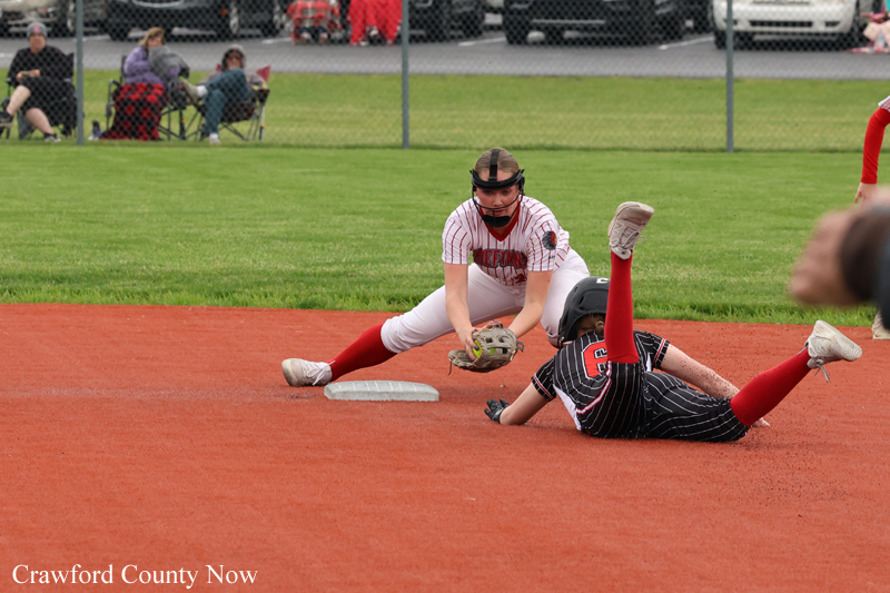 Softball infielder in white and red reaches for the ball near second base as a runner slides into the bag on a red-dirt field.