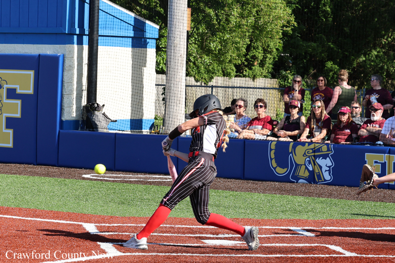 Softball player in black and red uniform swings at a pitch near home plate as spectators watch from blue dugout-style fencing.