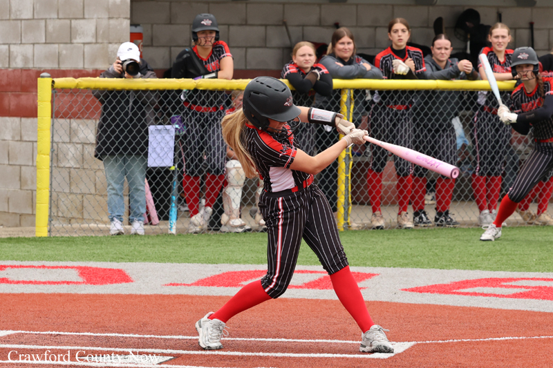 Female softball player in pinstriped uniform and red socks swings a pink bat at the plate, helmet on, during a game as teammates watch from the dugout behind a yellow fence.