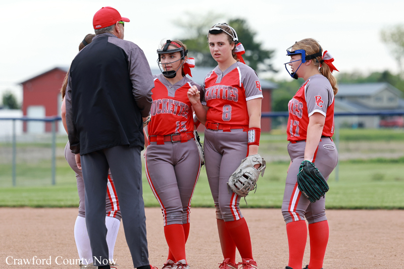 Youth softball team in red and gray uniforms listening to coach during a huddle on the infield.