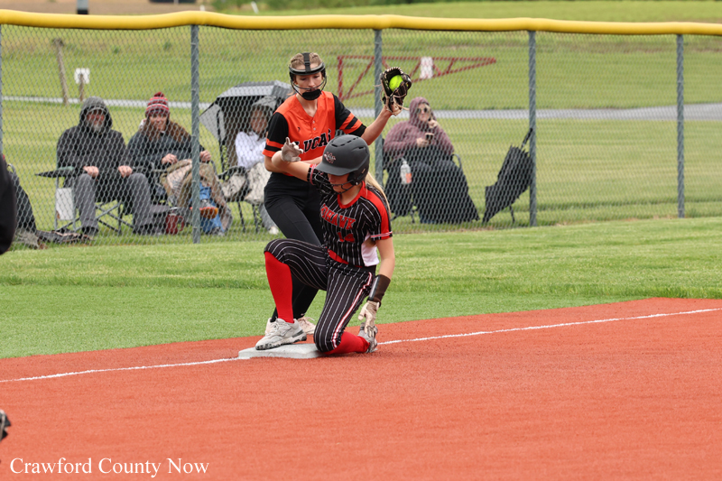 Softball runner slides into first base as the baseman covers the bag; orange and black uniforms, fans watching behind a chain-link fence.