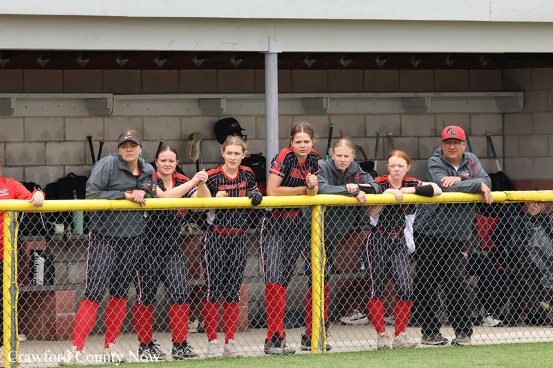 A group of young female softball players leaning on a yellow fence in the dugout, wearing black and red uniforms with red socks, watching something off-camera.