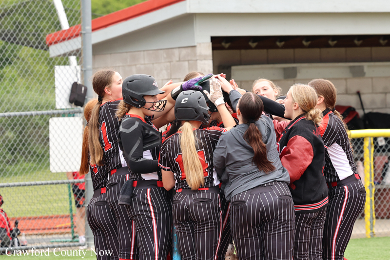 Softball team in pinstripe uniforms huddling together in the dugout for a pep talk before the game.