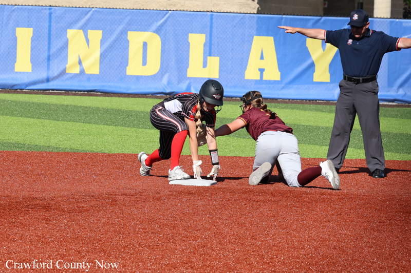Two softball players battle for a base as the runner slides while the fielder reaches for the bag, with an umpire signaling nearby.
