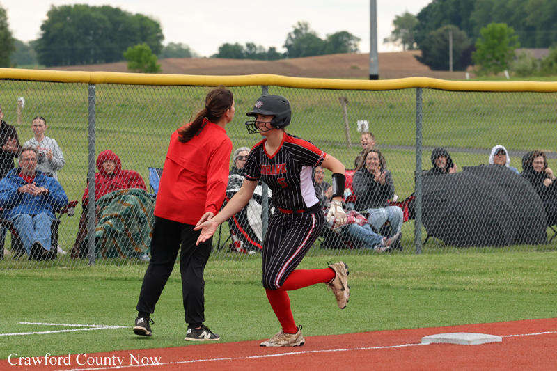 Girls softball player in a black pinstripe uniform with red socks runs toward home plate, greeted by a coach in red as spectators watch behind the fence.