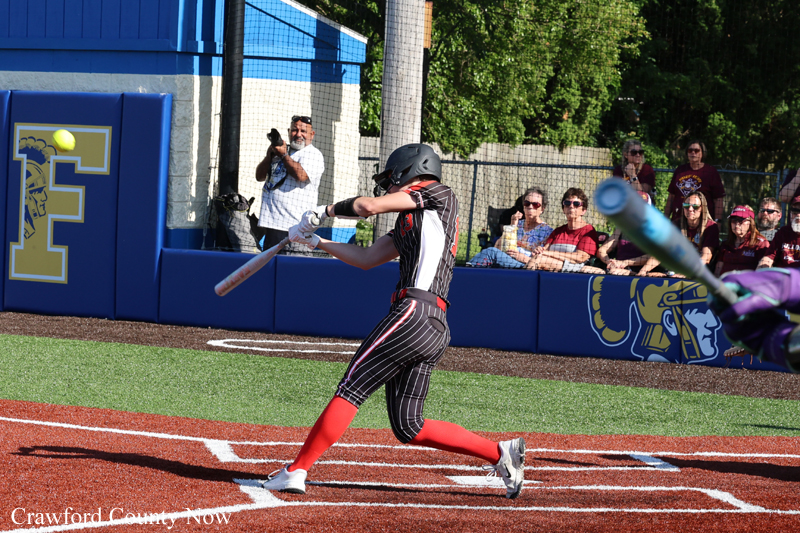 Softball batter in a black and red uniform swings at a pitched ball, with spectators and a blue padded wall behind her.