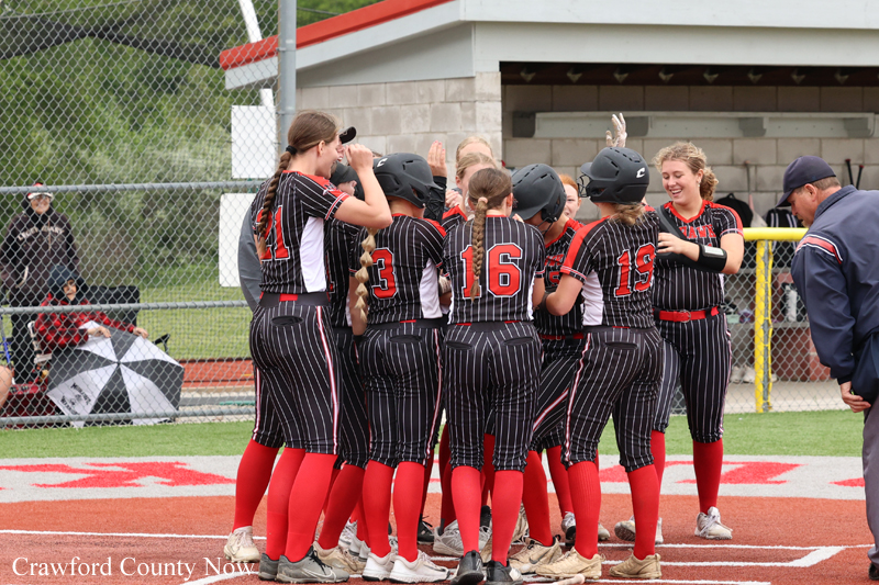 Girls softball team huddling in a circle on the field, wearing black pinstripe uniforms with red socks and helmets.