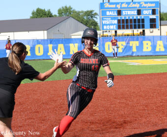 Softball player in a black pinstripe uniform high-fives a teammate near home plate; helmet on, red socks, with a scoreboard and blue outfield wall in the background.