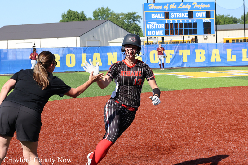 Softball player in a black pinstripe uniform high-fives a teammate near home plate; helmet on, red socks, with a scoreboard and blue outfield wall in the background.
