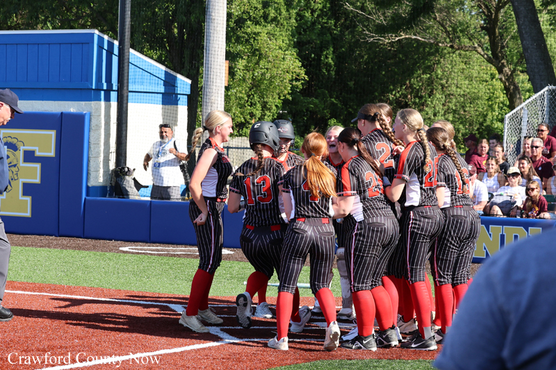 Softball team in black pinstripe uniforms gathers in a huddle on the field, wearing red socks and helmets, with fans watching in the background.