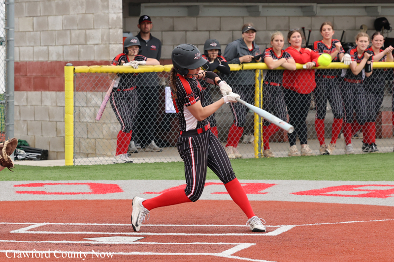 Female softball batter in black-red uniform swings at a pitch on an orange infield, helmet on, spectators behind a chain-link fence watching.