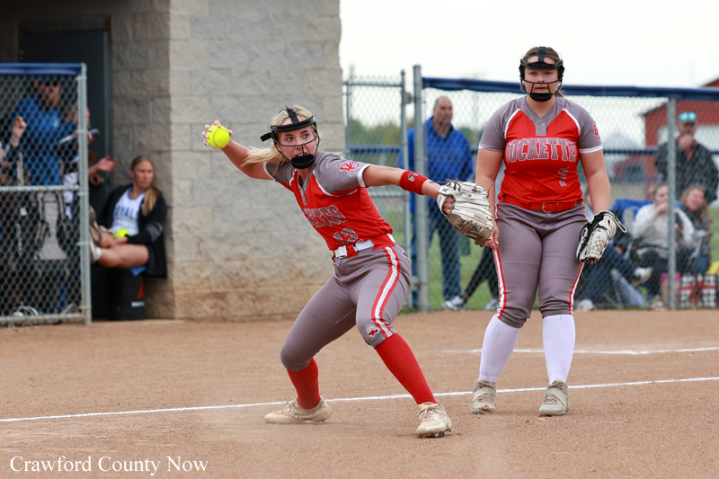 Softball pitcher in red-gray uniform winds up a throw on a dirt field, with catcher standing behind ready at home plate.