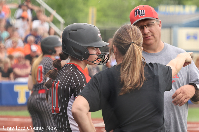 Softball player in a black helmet and pinstriped jersey (#11) listens to a coach pointing during a game, with spectators in the stands behind them.