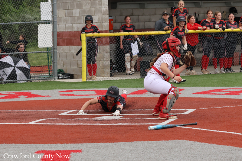 Softball scene at home plate: a baserunner slides headfirst while the catcher in red protective gear prepares at the plate, with teammates and spectators in the dugout area behind them.