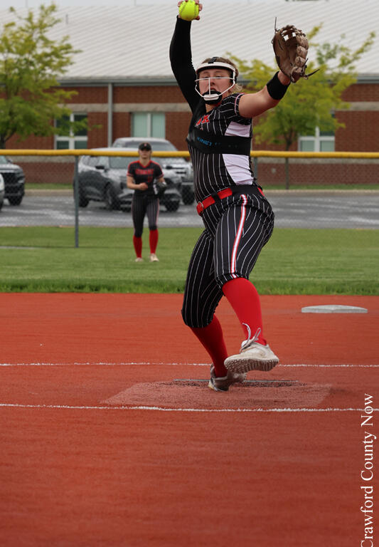 Softball pitcher in black pinstriped uniform throws from the mound, with glove raised and a bright green ball in hand; field and opposing player in background.