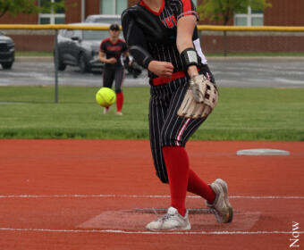 Softball pitcher in black-red uniform winds up on a red clay field, ball just left her hand with a catcher in the background.
