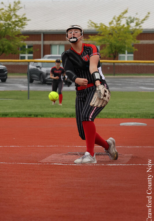 Softball pitcher in black-red uniform winds up on a red clay field, ball just left her hand with a catcher in the background.