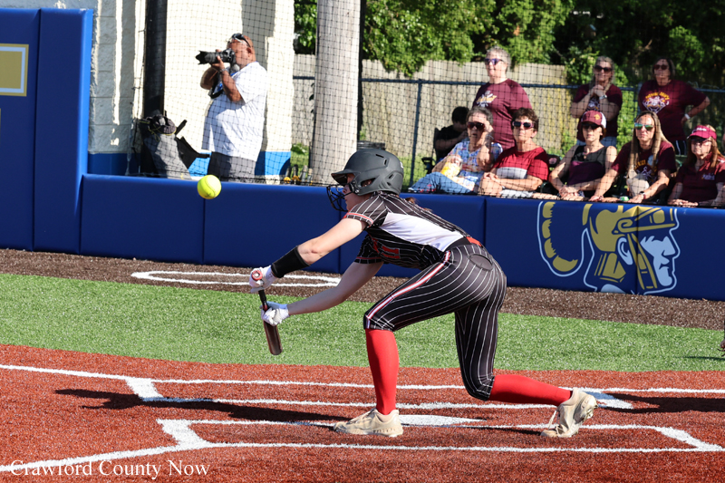 Female softball player in a black pinstripe uniform and red socks, wearing a helmet, reaching with her bat toward a pitched ball at home plate as spectators watch from the outfield wall.