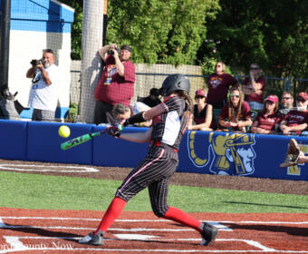 Softball player in a black pinstripe uniform with red socks swings at a pitch near home plate, while spectators watch from behind a blue outfield wall.
