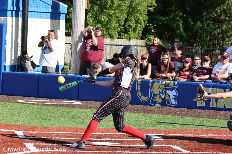 Softball player in a black pinstripe uniform with red socks swings at a pitch near home plate, while spectators watch from behind a blue outfield wall.