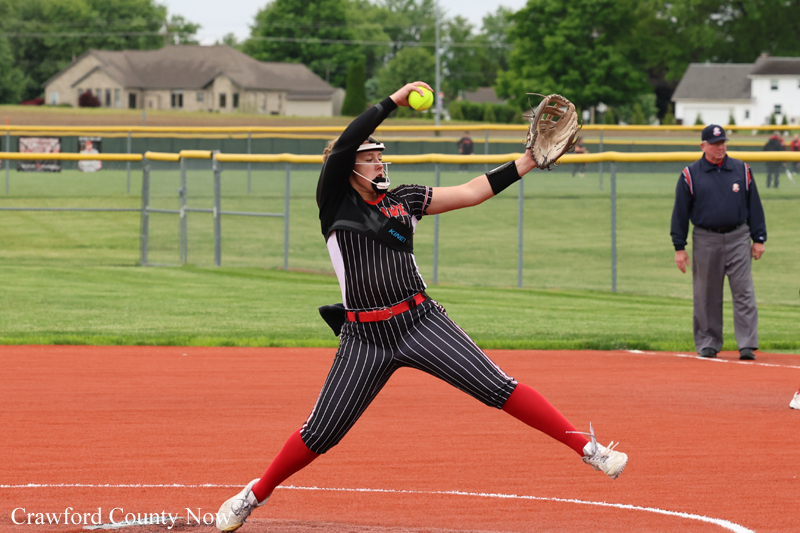 Softball pitcher in a pinstripe uniform winds up to throw on a red dirt mound, glove up and ball in hand.