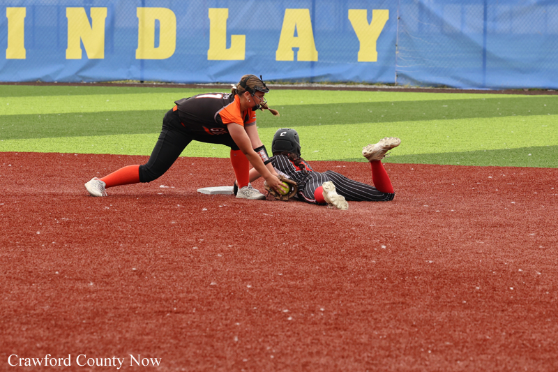 Fielder in orange and black tags a sliding baserunner at a base on a red dirt softball infield, with a blue outfield fence in the background.
