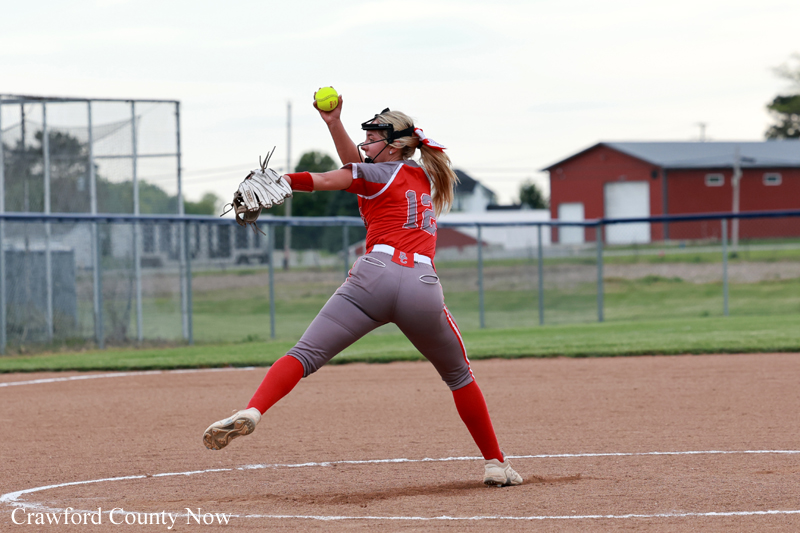 Softball pitcher in a red uniform mid-pitch on the mound, glove outstretched as she throws the ball.