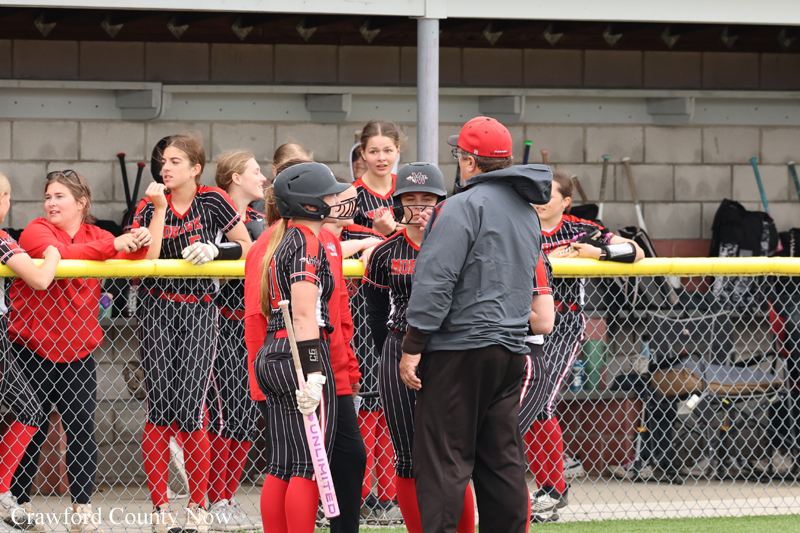 Softball players in black-and-red uniforms listen to a coach in a red cap near a chain-link fence at the dugout.