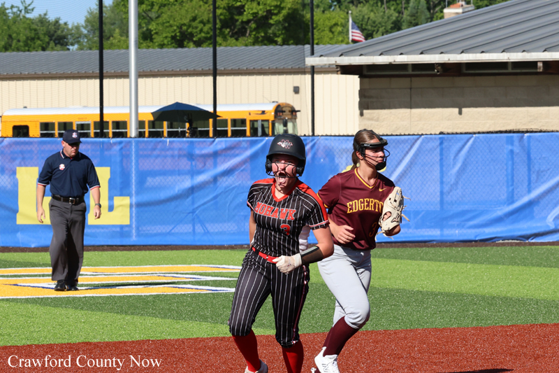 Two softball players sprint toward the base as an umpire watches in the background on a sunny field, one in black pinstripe with a helmet and the other in maroon with a glove behind her