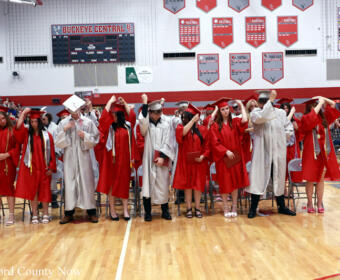 Group of graduates in red gowns (some wearing caps) and faculty in white robes stand in a gymnasium, preparing for a graduation ceremony with spectators in the stands and red banners on the walls.