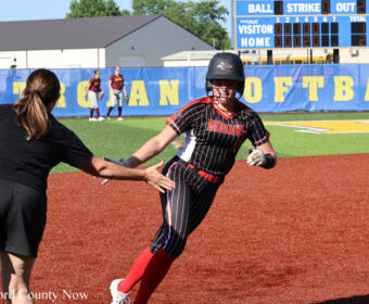 Softball baserunner in a black pinstripe uniform slides toward home while a teammate in black reaches out to assist on the dirt infield, blue outfield wall in the background.