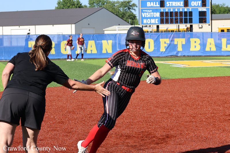 Softball baserunner in a black pinstripe uniform slides toward home while a teammate in black reaches out to assist on the dirt infield, blue outfield wall in the background.