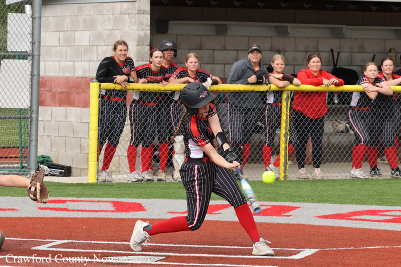 Female softball batter in a black-red uniform swings at a pitched ball near home plate, teammates watching from behind the fence, helmet on.
