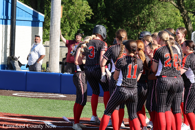 Girls' softball team in black pinstripe uniforms with red numbers gather in a huddle on the field as a coach or staff member speaks near the dugout.