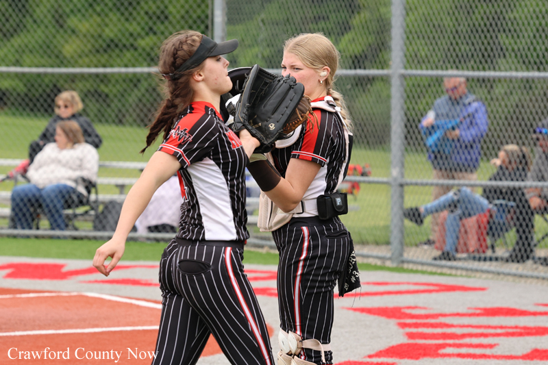 Two female softball players in black pinstripe uniforms celebrate at home plate; one is the catcher with gear and glove.