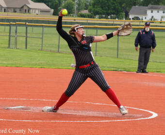 Female softball pitcher in a black pinstripe uniform with red socks delivering a pitch from the mound, yellow ball in hand, coach watching in the background.