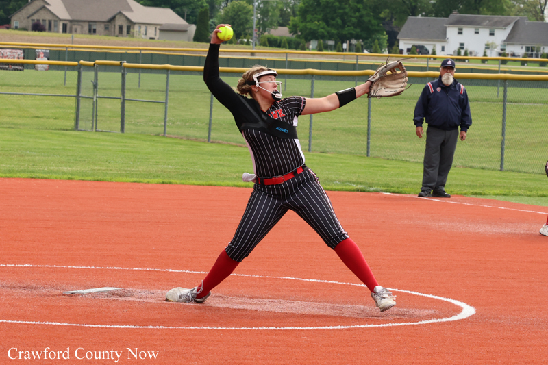 Female softball pitcher in a black pinstripe uniform with red socks delivering a pitch from the mound, yellow ball in hand, coach watching in the background.
