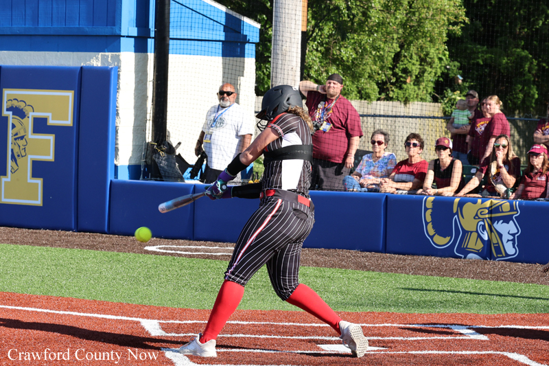 Female softball player in black pinstripe uniform and red socks swings at a pitch near home plate as spectators watch from the blue dugout wall behind her.