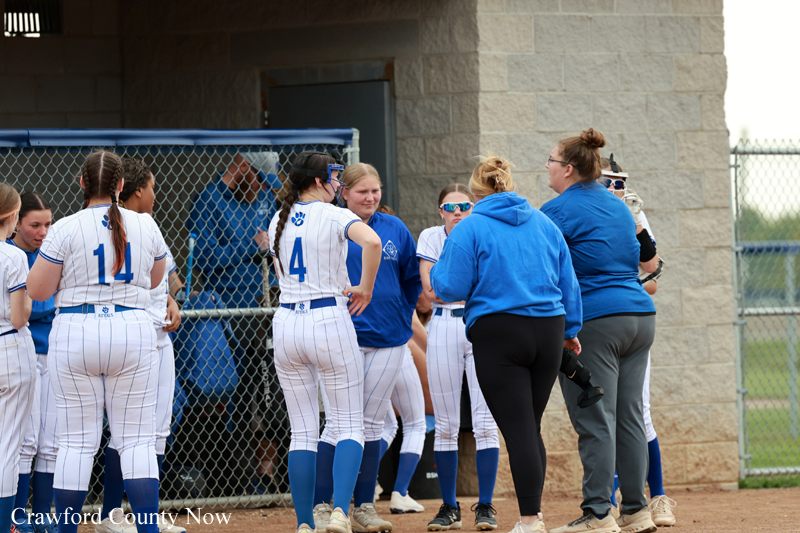 Girls' softball team huddling near the dugout with coaches in blue hoodies on a dirt field fence backdrop.