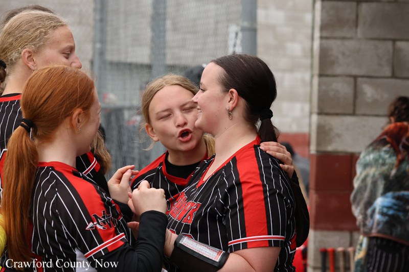 Group of female softball players in black-and-red striped uniforms share a hug, with one woman kissing another on the cheek in a celebratory moment.