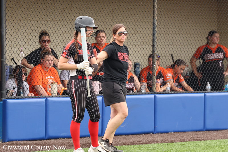 Female softball batter stands at the plate in uniform with helmet, ready to bat, while a coach in black shirt watches nearby from the dugout.