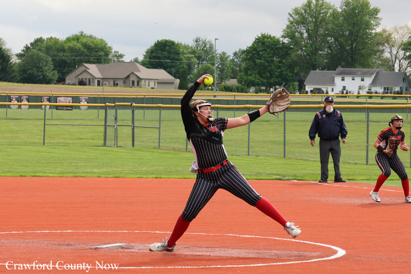 Female softball pitcher in a black pinstripe uniform delivers a pitch from the mound on an orange dirt field, with a teammate and umpire in the background.