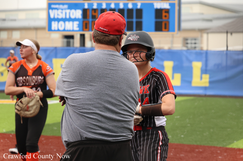 Coach in gray shirt and red cap talks to a young softball player in a helmet on the field, with the scoreboard in the background.