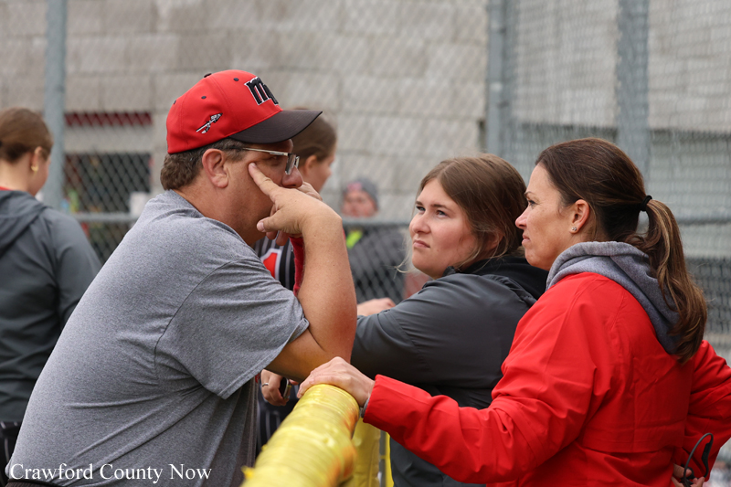 Three spectators leaning on a yellow railing beside a chain-link fence, chatting at a sports venue.