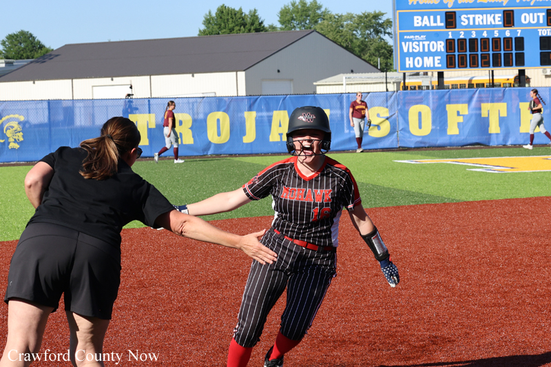 Softball player in a black pinstripe uniform with helmet high-fives a coach on the red infield as the game continues behind them.