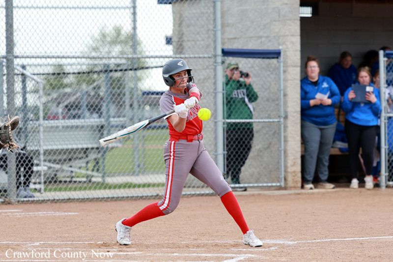 Female softball batter in red and gray swings at a pitch during a game, near a chain-link fence and dugout with teammates watching in blue jackets.