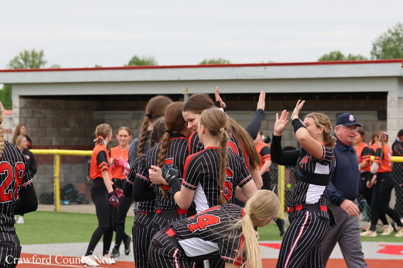 Girls' softball teammates in black and red uniforms embrace in a congratulatory group hug on the field near the dugout, with a coach watching nearby.