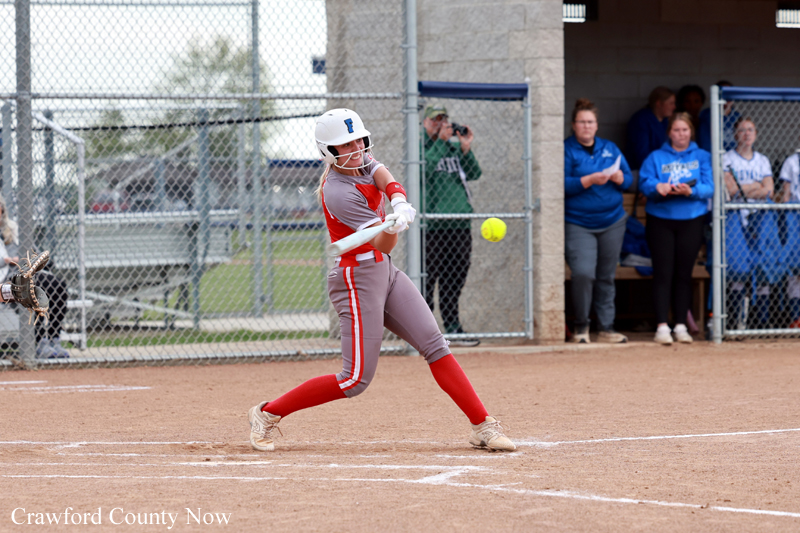 Female softball batter in a red-gray uniform swings at a pitch on a dirt field, helmet on, with dugout and spectators in the background.
